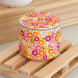 Peut inclure: Un récipient cylindrique peint à la main avec un couvercle, orné d'un motif de cercles colorés dans les tons de rose, orange, jaune et or. Le récipient est posé sur une petite palette en bois. Le design est un mélange d'éléments abstraits et décoratifs.