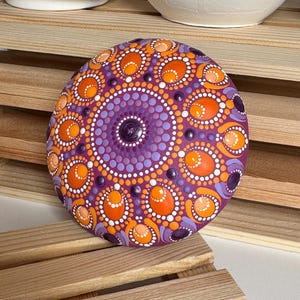 Handbemalter Mandala-Stein mit magentafarbener Grundierung und Dot Painting in Orange, Apricot, Lila und Flieder. Die Aufnahme zeigt das harmonische Zusammenspiel der warmen und kühlen Farbtöne sowie die charakteristischen ovalen Mandala-Elemente. Erhabene Farbpunkte und feine Details verleihen dem Kunstwerk Tiefe und eine besondere handgefertigte Ausstrahlung.
