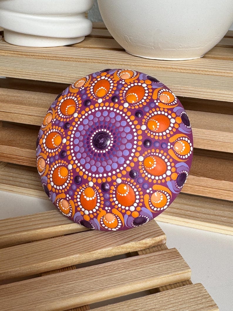 Handbemalter Mandala-Stein mit magentafarbener Grundierung und Dot Painting in Orange, Apricot, Lila und Flieder. Die Aufnahme zeigt das harmonische Zusammenspiel der warmen und kühlen Farbtöne sowie die charakteristischen ovalen Mandala-Elemente. Erhabene Farbpunkte und feine Details verleihen dem Kunstwerk Tiefe und eine besondere handgefertigte Ausstrahlung.