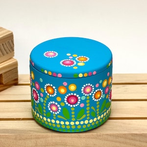 Könnte beinhalten: Eine runde, türkisfarbene Box mit Deckel, verziert mit einem Blumenmuster aus rosa, orangefarbenen, gelben und weißen Punkten. Die Box steht auf einer Holzoberfläche. Durchmesser ca. 10 cm.