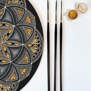 Può includere: Un'opera d'arte mandala nera con dettagli a punti dorati, grigi e bianchi. Sono visibili anche tre sottili pennelli neri e un piccolo barattolo di vernice dorata, che suggeriscono un progetto artigianale o artistico.