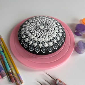 Könnte beinhalten: Ein schwarzer Stein mit einem darauf gemalten weißen und grauen Mandala-Muster. Der Stein liegt auf einem rosa Drehteller. Auf dem Tisch liegen mehrere Pinsel und Farbflaschen.