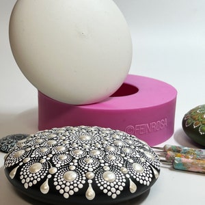 Peut inclure: Un rocher peint en noir et blanc avec un motif de mandala. Le rocher est posé sur une surface blanche à côté d'un moule en silicone rose et d'un rocher blanc. Le moule porte le texte "FEINROSA" sur le côté.