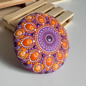 Handbemalter Mandala-Stein als dekoratives Kunstobjekt auf einer Holzablage im Wohnbereich. Das Dot Painting in Orange, Apricot, Lila und Flieder hebt sich kontrastreich von der magentafarbenen Grundierung ab. Die symmetrische Mandala-Gestaltung mit ovalen und runden Punktformen verleiht dem Unikat eine harmonische Ausstrahlung und macht es zu einem besonderen Blickfang für Wohnräume und kreative Rückzugsorte.