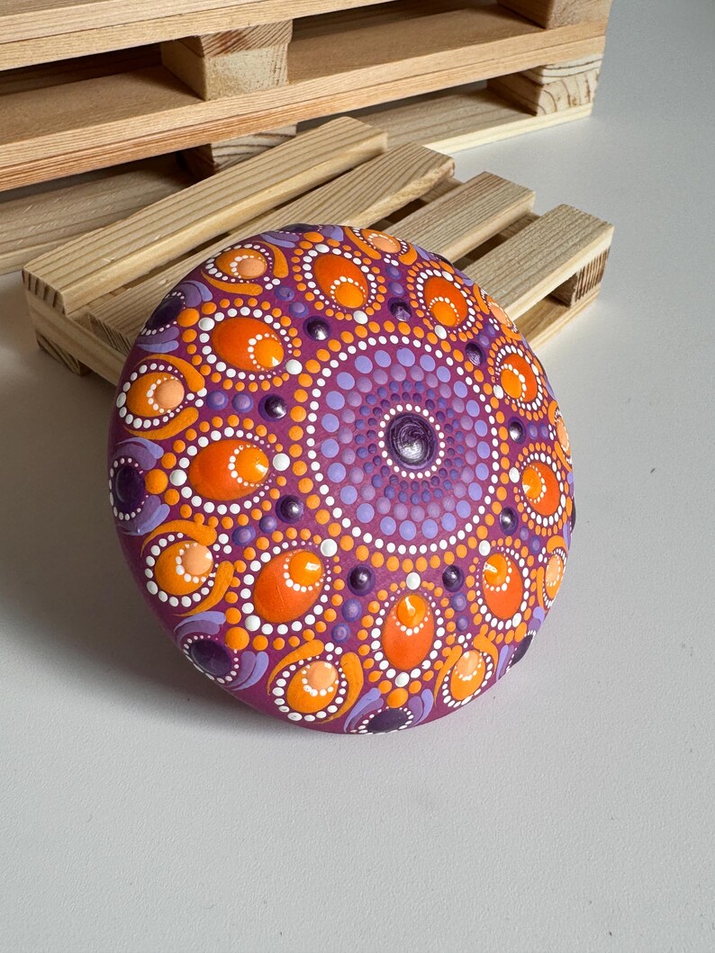 Handbemalter Mandala-Stein als dekoratives Kunstobjekt auf einer Holzablage im Wohnbereich. Das Dot Painting in Orange, Apricot, Lila und Flieder hebt sich kontrastreich von der magentafarbenen Grundierung ab. Die symmetrische Mandala-Gestaltung mit ovalen und runden Punktformen verleiht dem Unikat eine harmonische Ausstrahlung und macht es zu einem besonderen Blickfang für Wohnräume und kreative Rückzugsorte.