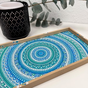 Könnte beinhalten: Ein Holztablett mit einem blauen und grünen Mandala-Design, das auf den Boden gemalt ist. Das Mandala besteht aus kleinen Punkten in verschiedenen Blautönen und Grüntönen.