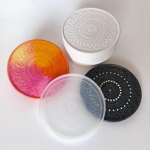 Può includere: Un set di sottobicchieri decorativi in vari colori e design. Include una pila di sottobicchieri bianchi con un motivo geometrico in rilievo, un sottobicchiere nero con pois bianchi e un sottobicchiere trasparente. L'altro ha un gradiente arancione e rosa.