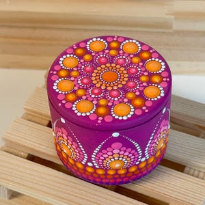 Può includere: Una piccola scatola rotonda viola con un elaborato design a mandala sul coperchio e sui lati. Il mandala presenta motivi a pois arancioni, rosa e bianchi. La scatola è appoggiata su un piccolo pallet di legno.