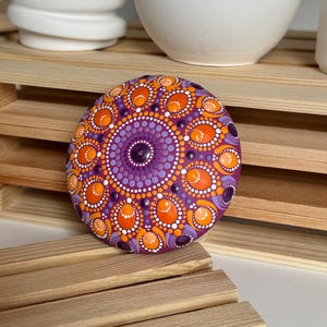 Gesamtansicht eines handbemalten Mandala-Steins auf einer Holzablage. Das Dot Painting in Orange, Apricot, Lila und Flieder bildet ein symmetrisches Mandala auf magentafarbenem Untergrund. Die Aufnahme zeigt die Größenwirkung des Kunstobjekts im Wohnbereich sowie die ausgewogene Farbkomposition aus warmen und kühlen Farbtönen. Ovale und runde Punktformen verleihen dem Mandala eine besondere Tiefe und Struktur.