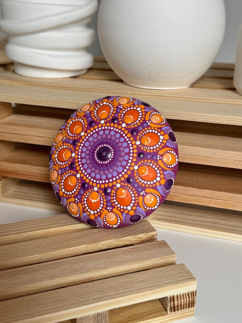 Gesamtansicht eines handbemalten Mandala-Steins auf einer Holzablage. Das Dot Painting in Orange, Apricot, Lila und Flieder bildet ein symmetrisches Mandala auf magentafarbenem Untergrund. Die Aufnahme zeigt die Größenwirkung des Kunstobjekts im Wohnbereich sowie die ausgewogene Farbkomposition aus warmen und kühlen Farbtönen. Ovale und runde Punktformen verleihen dem Mandala eine besondere Tiefe und Struktur.