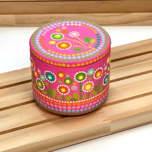 Può includere: Una scatola decorativa rotonda rosa con coperchio, ornata da un colorato motivo floreale. La scatola presenta un design vibrante di fiori e punti nei toni del verde, giallo, arancione e blu. La scatola è posta su una superficie di legno.