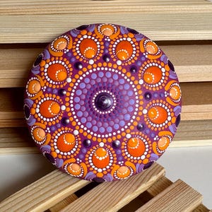 Frontalansicht eines handbemalten Mandala-Steins mit magentafarbener Grundierung. Das symmetrische Dot Painting zeigt detailreiche Muster in Orange, Apricot, Lila und Flieder. Runde und ovale Punkte bilden konzentrische Mandala-Kreise mit erhabenen Strukturen. Der Stein steht dekorativ auf einer Holzablage und zeigt die harmonische Farbwirkung des Kunstwerks besonders deutlich.
