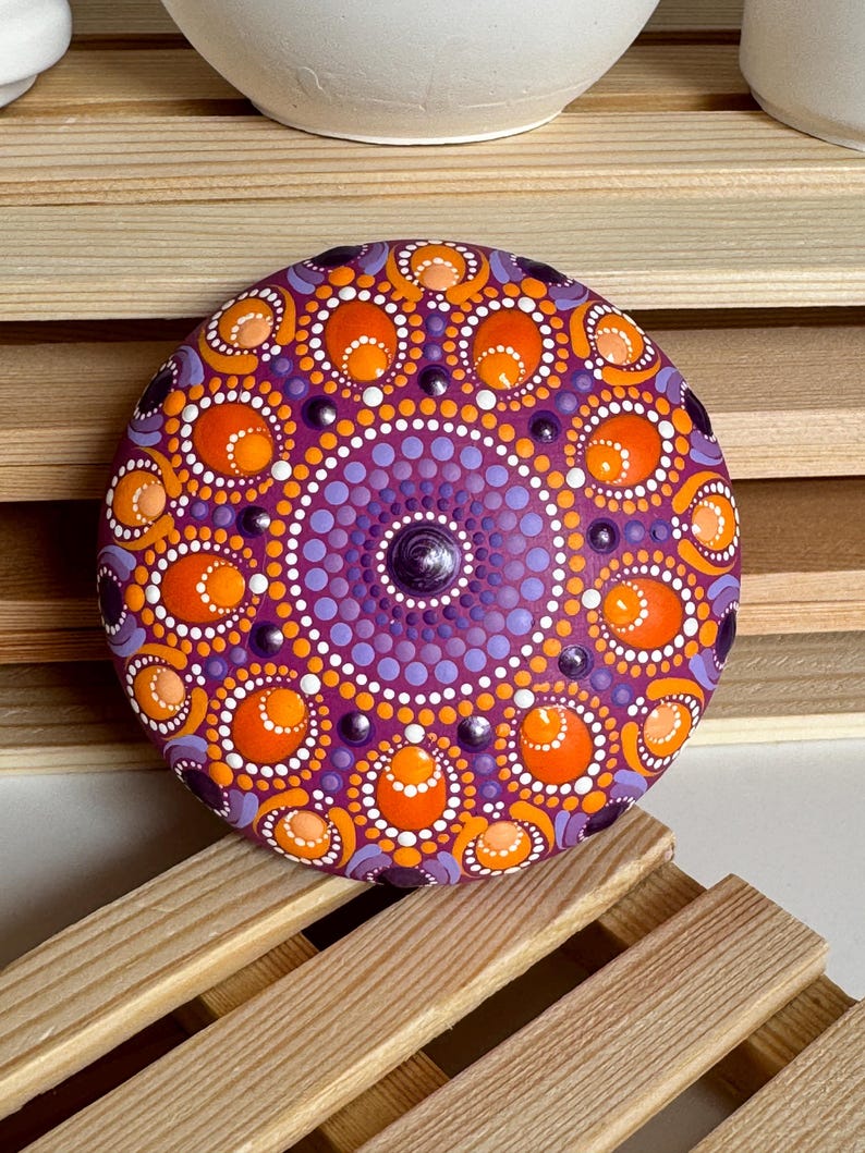 Frontalansicht eines handbemalten Mandala-Steins mit magentafarbener Grundierung. Das symmetrische Dot Painting zeigt detailreiche Muster in Orange, Apricot, Lila und Flieder. Runde und ovale Punkte bilden konzentrische Mandala-Kreise mit erhabenen Strukturen. Der Stein steht dekorativ auf einer Holzablage und zeigt die harmonische Farbwirkung des Kunstwerks besonders deutlich.