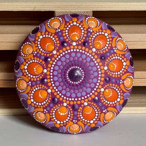 Frontalansicht eines handbemalten Mandala-Steins mit symmetrischem Dot Painting auf magentafarbenem Untergrund. Das Mandala kombiniert Orange, Apricot, Lila und Flieder zu einem harmonischen Farbspiel. Die Aufnahme zeigt die vollständige Gestaltung mit runden und ovalen Punktformen, die sorgfältig von Hand aufgetragen wurden und dem Kunstwerk eine besondere Struktur und Tiefe verleihen.