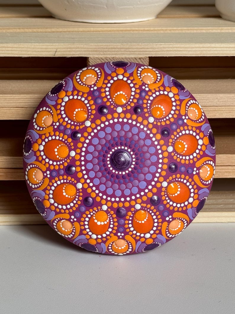 Frontalansicht eines handbemalten Mandala-Steins mit symmetrischem Dot Painting auf magentafarbenem Untergrund. Das Mandala kombiniert Orange, Apricot, Lila und Flieder zu einem harmonischen Farbspiel. Die Aufnahme zeigt die vollständige Gestaltung mit runden und ovalen Punktformen, die sorgfältig von Hand aufgetragen wurden und dem Kunstwerk eine besondere Struktur und Tiefe verleihen.