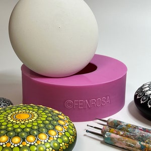 Könnte beinhalten: Eine rosa Silikonform zum Erstellen von runden, glatten, dekorativen Steinen. Die Form ist mit einer weißen Kugel im Inneren dargestellt, sowie zwei bemalten Steinen mit Mandala-Designs. Die Steine sind grün, gelb und weiß sowie schwarz und weiß. Das Bild zeigt auch einen Satz von Punktwerkzeugen zum Malen.