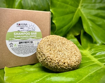 May include: A round, brown shampoo bar with a rough texture, labeled "KAHUNA PRODUCTS" and "SHAMPOO BAR YLANG-YLANG & JASMINE", sitting on a green leaf.