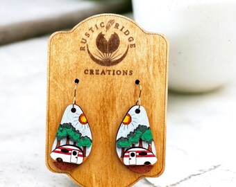 May include: A pair of wooden earrings with a camper van, trees, and a sun painted on them. The earrings are on a wooden display card with the text "Rustic Ridge Creations".