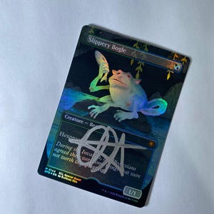 May include: A holographic trading card featuring the creature "Slippery Bogle." The card has a colorful illustration of a frog-like creature and a fish. The card is signed with a silver marker. The text "Slippery Bogle" is at the top.