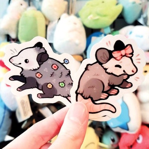 May include: Two illustrated stickers of opossums. One opossum is gray with a floral pattern and the other is brown with a pink bow. Both opossums are smiling.