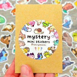 May include: A rectangular brown paper envelope featuring a circular white sticker with colourful cartoon animal illustrations. The text reads mystery mini stickers @spicymochi with three question marks in red, blue, and yellow.