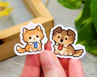 Puede incluir: Dos pegatinas troqueladas con un gato atigrado naranja y un perro marrón y blanco de estilo dibujo animado. Ambos animales están sentados, llevan camisas blancas con cuello y corbatas, una azul y otra naranja, con un borde blanco.
