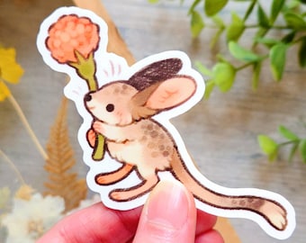 May include: A die-cut sticker featuring a cartoon jerboa holding a flower. The jerboa has tan fur with brown markings and large ears. The flower is orange, and the sticker has a white border. The sticker is held by a hand.