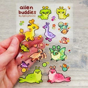 May include: A sheet of clear stickers with cartoon alien characters in various colours. The stickers feature creatures in green, yellow, pink, and purple, with the text "alien buddies by Spicymochi" at the top.