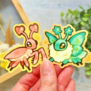 May include: Two colorful stickers featuring whimsical insect designs. One is a pink mantis with heart-shaped antennae, and the other is a teal and yellow insect with flower-like antennae and wings. Both are on a yellow background.
