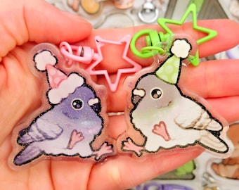 May include: Two clear acrylic keychains featuring cartoon pigeons wearing party hats. One pigeon is purple with a pink hat, and the other is gray with a green hat. Both have pink feet and are attached to star-shaped clasps.
