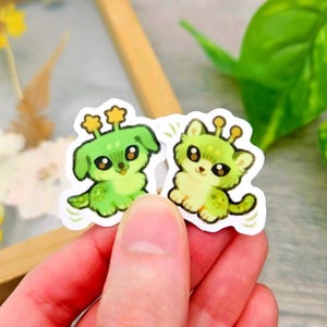 May include: Two stickers depicting adorable green cartoon creatures with large eyes and star-shaped antennae. One is dog-like, the other cat-like. The stickers have a white border and are held in a hand.