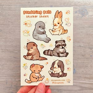 May include: A sticker sheet titled Pondering Pals featuring six illustrated animals including a gray seal, a tan rabbit, a brown otter, a raccoon, an orange tabby kitten, and a lop-eared rabbit, all surrounded by small colorful cloud and star icons.