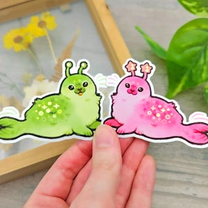 May include: Two stickers featuring cartoon creatures. One is lime green with white and yellow accents, the other is pink with white and pink accents. Both have antennae and are in a lying down pose.