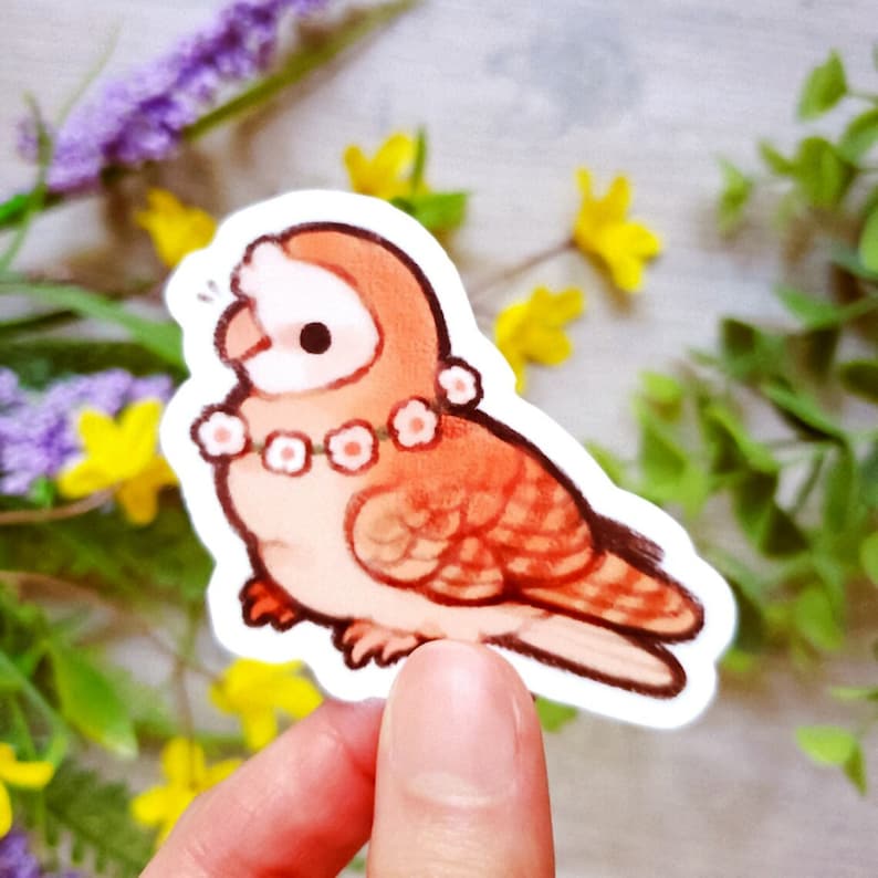 May include: A sticker depicting a cartoon bird with orange and white plumage, adorned with a flower lei. The bird has a rounded form and is positioned on a surface. The background features flowers and foliage.