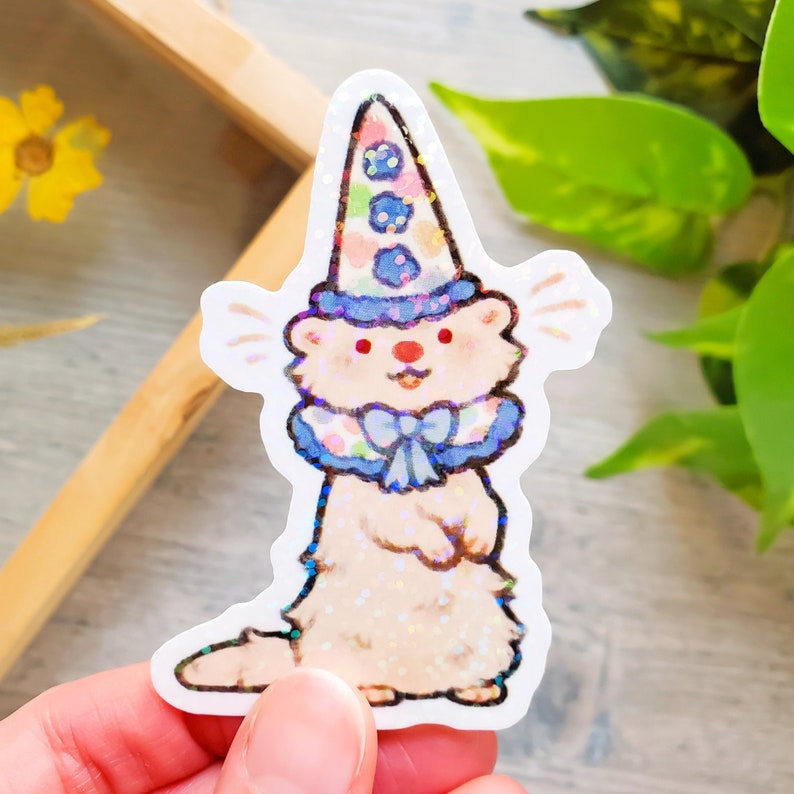 May include: A white ferret wearing a blue and pink clown hat with a blue bow tie. The ferret is standing on its hind legs and has a surprised expression on its face. The background is a white surface.