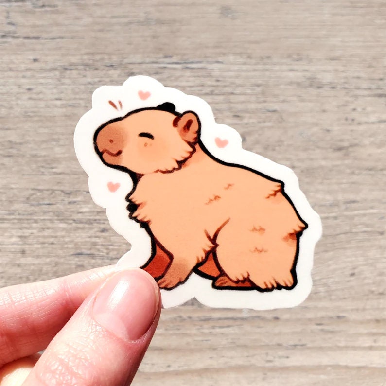 May include: A sticker of a cartoon capybara with a pink blush and pink hearts around it. The capybara is sitting on its hind legs and has a happy expression.