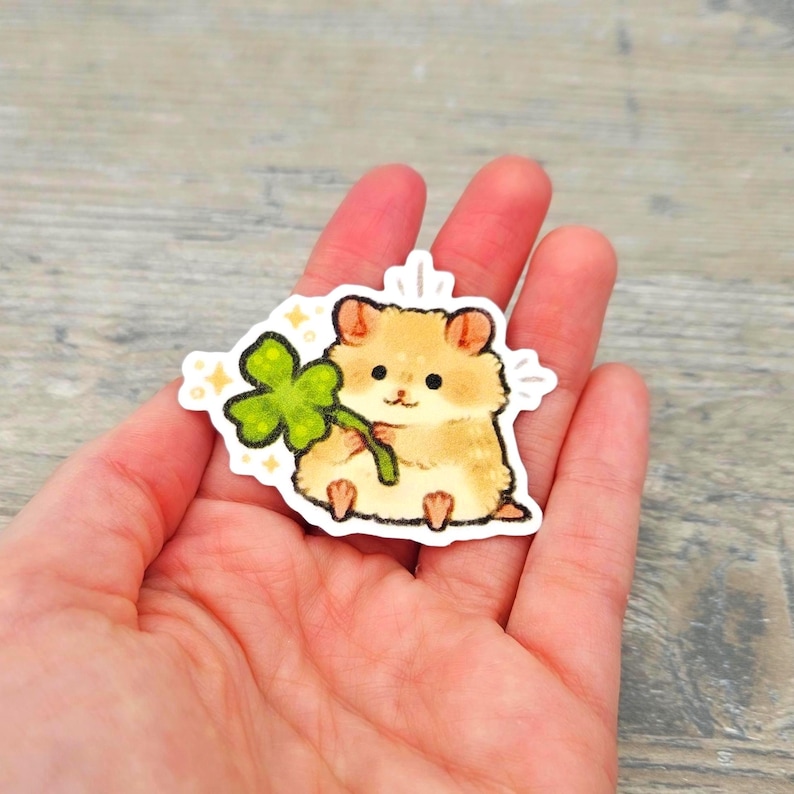 May include: A die-cut sticker featuring a light brown hamster with dark eyes and small ears, holding a bright green four-leaf clover. The illustration has a soft, textured appearance with small yellow star accents and a white border outline.