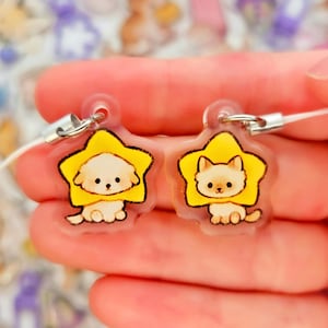 May include: Two clear, star-shaped charms featuring cartoon animals. One charm has a white dog, the other a tan cat, both wearing yellow star-shaped frames. Each charm has a silver-colored clasp and a white cord.