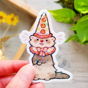 May include: A sparkly sticker of a cartoon ferret wearing a colorful party hat and a pink bow. The ferret is standing on its hind legs with its tail curled behind it.
