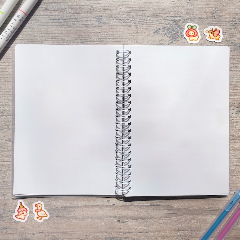 Puede incluir: Un cuaderno blanco de espiral con p&aacute;ginas en blanco. El cuaderno est&aacute; abierto y acostado sobre una superficie de madera. Hay dos marcadores y dos pegatinas en la superficie.