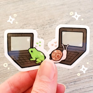 May include: Two laptop stickers featuring a frog and a snail. The frog is green and sitting on a black laptop with a white screen. The snail is brown and sitting on a black laptop with a white screen. Both stickers have a white background.