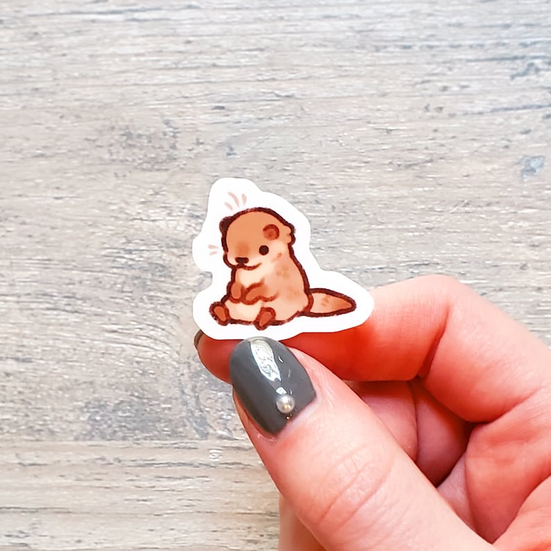 May include: A small, brown and white cartoon otter sticker with a surprised expression. The sticker is on a white background and is being held by a hand.