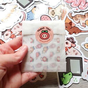 May include: A white paper bag with a pink and white sticker that says "Thank You! Spicymochi" on it. The bag is filled with small, colorful stickers featuring cartoon animals and other designs.