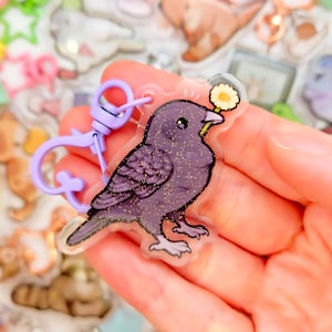 May include: A purple glittery bird keychain with a yellow flower in its beak and a light purple clasp. The bird is a cartoon illustration with black eyes and a white outline. Other similar keychains are in the background.