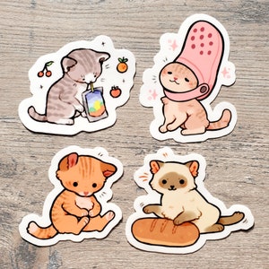 May include: Four sticker designs featuring cute cartoon cats in various poses. One cat is wearing a pink clog, another is drinking juice, one is sitting with its paws tucked in, and the last is playing with a baguette.