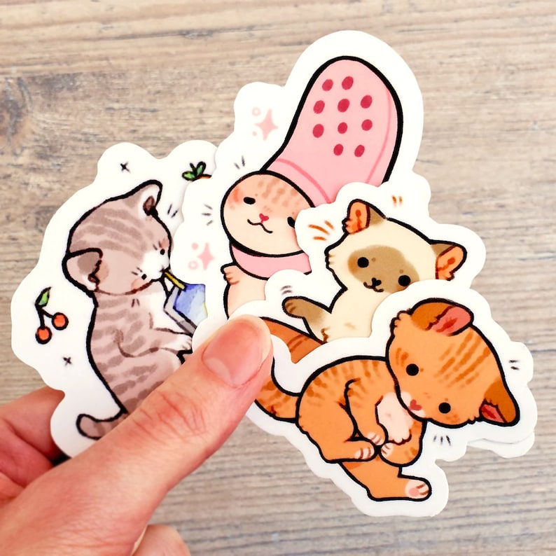 May include: Four colorful cat stickers with different poses and expressions. The stickers are on a light wood background. One cat is wearing a pink slipper on its head.