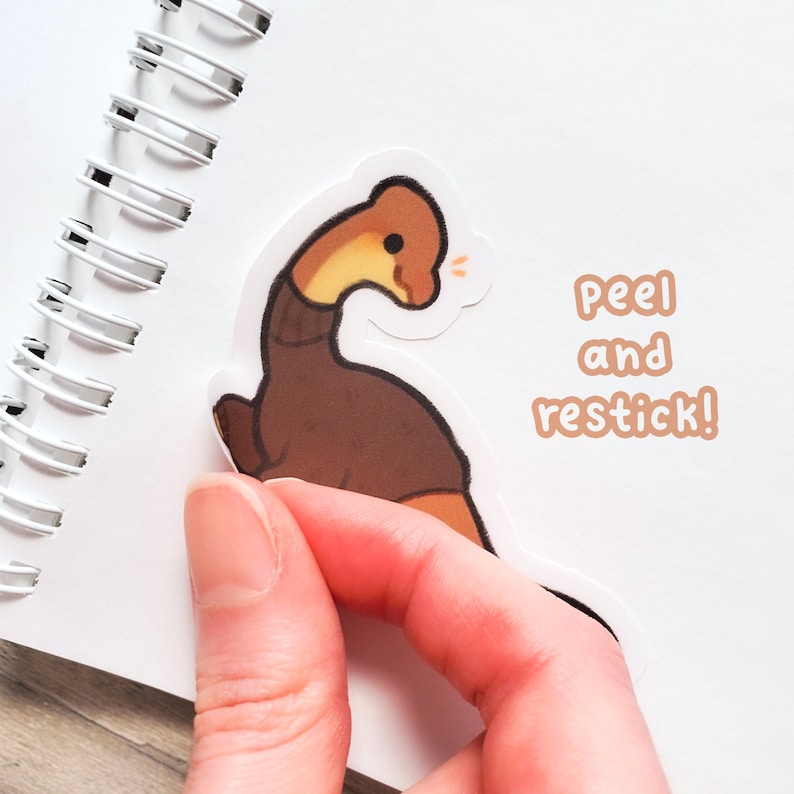 Puede incluir: Una pegatina de dibujos animados marr&oacute;n y tostado de un plesiosaurio con el texto "peel and restick!" sobre un fondo blanco.
