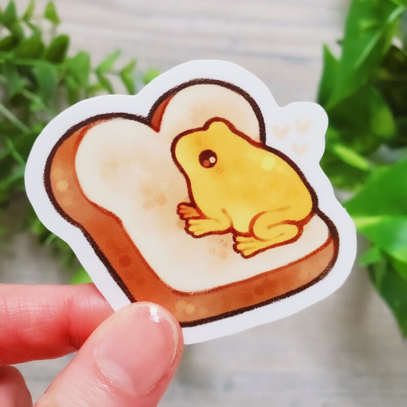 May include: A yellow frog sitting on a piece of toast. The frog is facing to the right and has a brown eye. The toast is brown and has a white center. There are three white hearts in the upper right corner of the image.