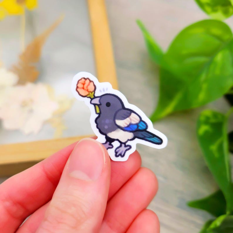 May include: A sticker featuring a cartoon magpie holding a small orange flower in its beak. The bird has a black head and back, with blue and white accents on its wings. The sticker is held by a hand.