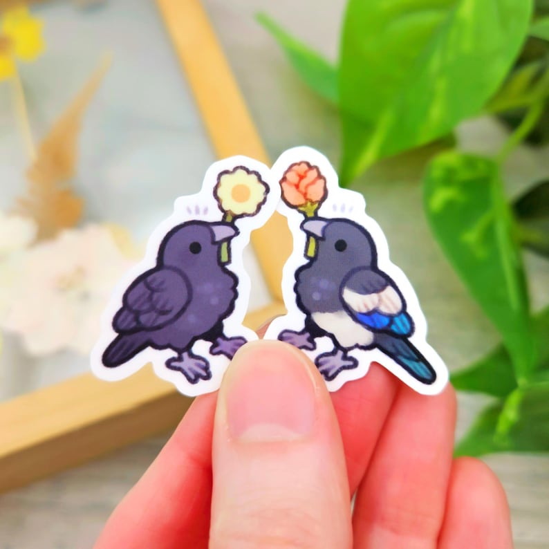 May include: Two bird stickers. One is a dark purple crow holding a yellow flower, and the other is a black and white magpie holding an orange flower. Both stickers have a white border and are held in a hand.